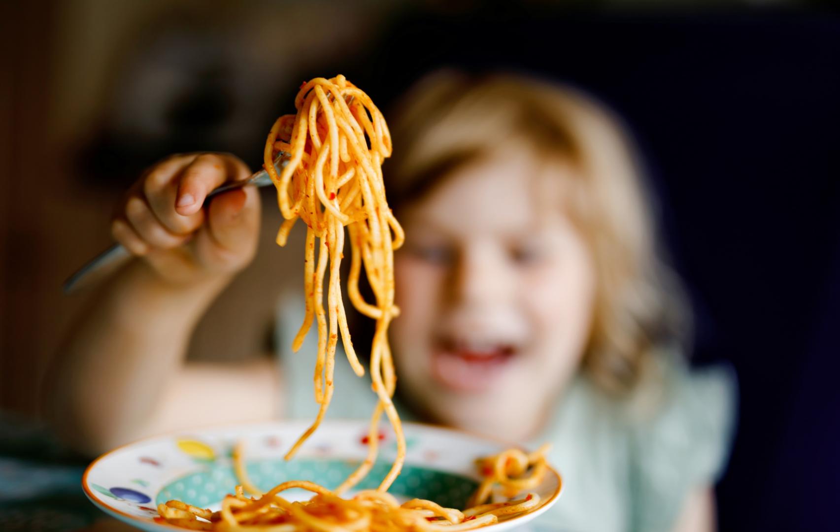 Bambina felice mangia spaghetti con una forchetta, sfondo sfocato.
