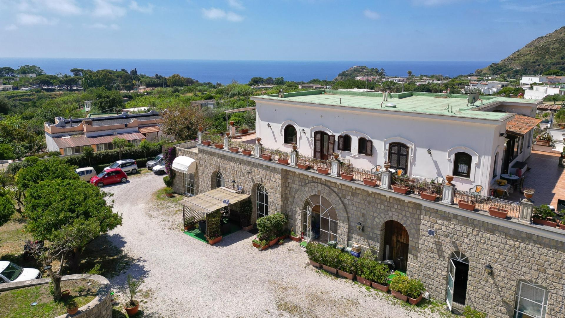 Villa mediterranea con vista mare, circondata da giardini e auto parcheggiate.