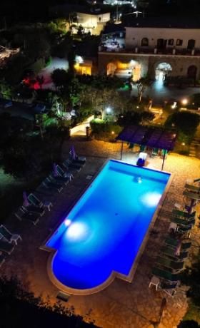 Piscina illuminata di notte con sedie a sdraio, circondata da un giardino e edifici.