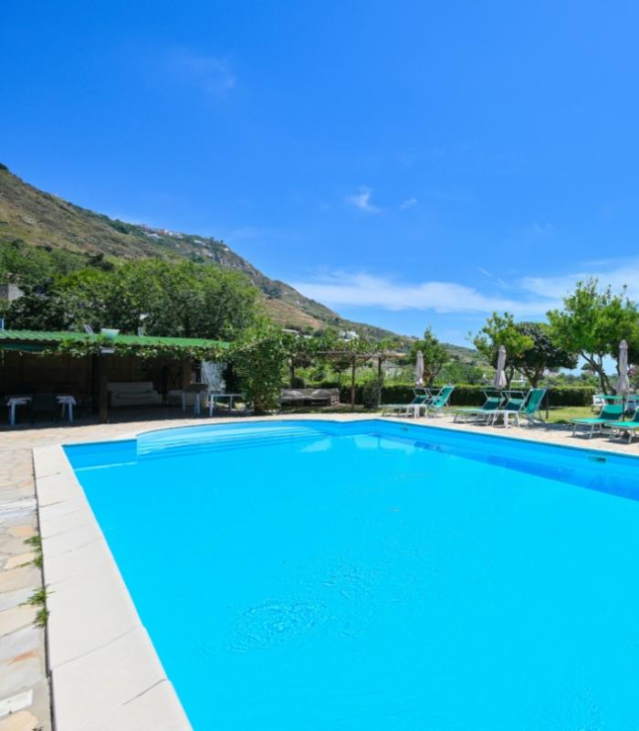 Piscina all'aperto con lettini, vista mare e colline, cielo limpido e soleggiato.