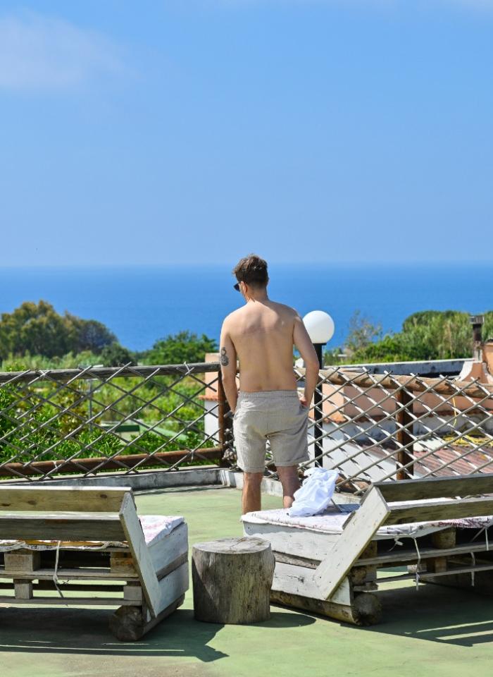 Uomo in terrazza con vista mare, sedie di legno e cielo azzurro.